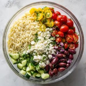 Mixing the Greek Orzo Salad ingredients in a bowl, with fresh tomatoes, cucumbers, olives, and feta